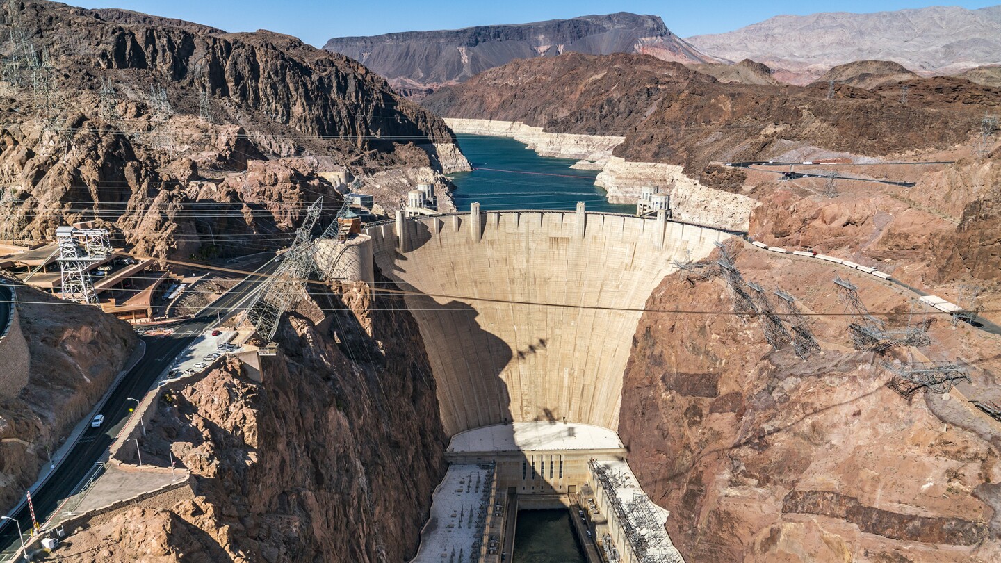 A wide image of the Hoover Dam, a concrete dam holding water from the Colorado River behind it. Surrounding the dam and river is a dry, red and brown mountainous landscape with electric wires crossing across the dam. 