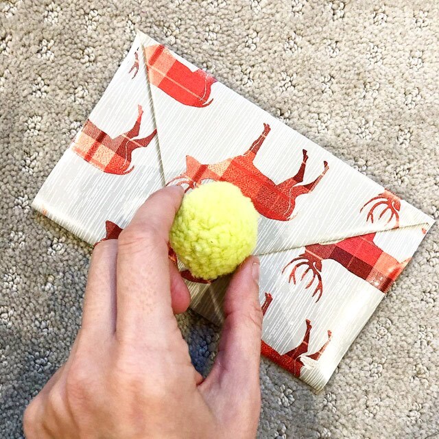 An envelope made out of wrapping paper decorated with a yellow pom pom.