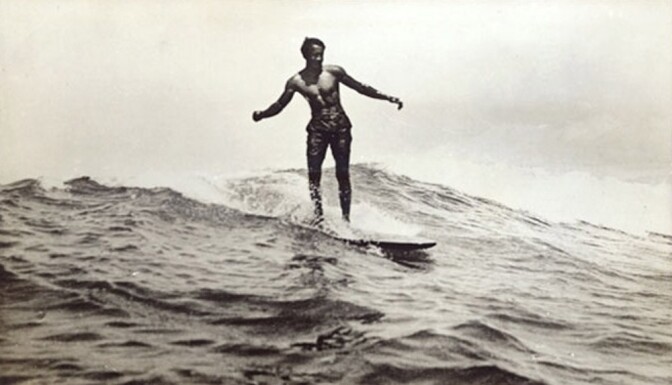 A black and white old photo of Duke Kahanamoku riding a wave in Waikiki. His arms are stretched out from his sides as he leans to the side slightly.