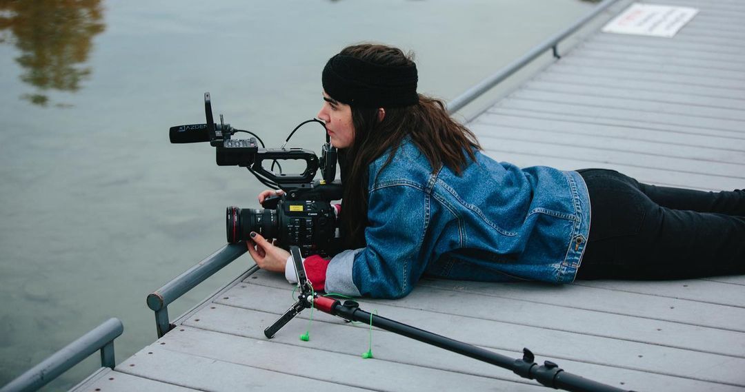 A woman in a denim blue jacket lays on her stomach on a dock. She's handling a camera that is pointed out towards a lake.