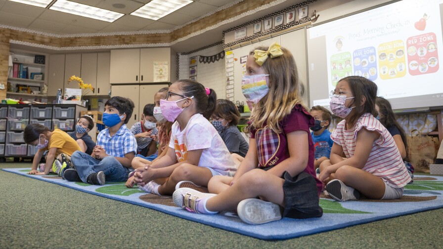 Children wearing masks sit on a rug in a classroom