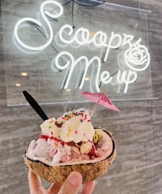 An ice cream sundae is served in half a coconut shell. The sundae is topped with rainbow sprinkles, a strawberry syrup drizzle and a pink paper umbrella. The sundae is held up against a wall that has a neon side that reads: "Scoop me up."