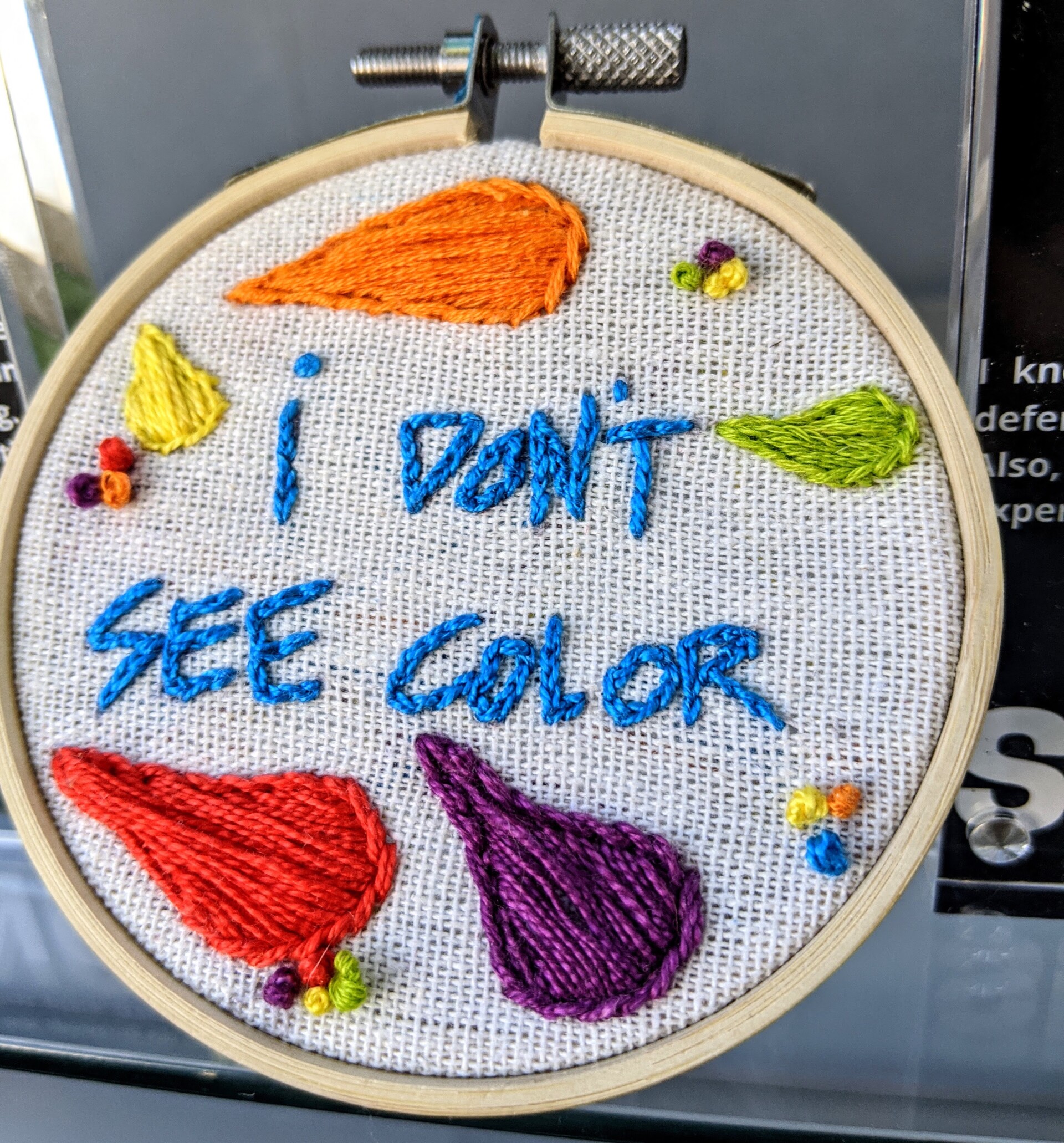 A small embroidery hoop with cream fabric stretched across embroidered with the words "I don't see color" in bright blue thread. Surrounding the words are teardrop shapes embroidered in various sizes and colors like red, orange, yellow, green and purple.