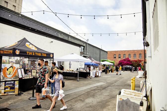 Masked and arm in arm, two attendees walk through the booths and tents at the FilLed Market in Downtown L.A. A nearby tent reads, "Tim's Thai Tea." The attendees are holding miscellaneous plastic bags and a bag with drinks.