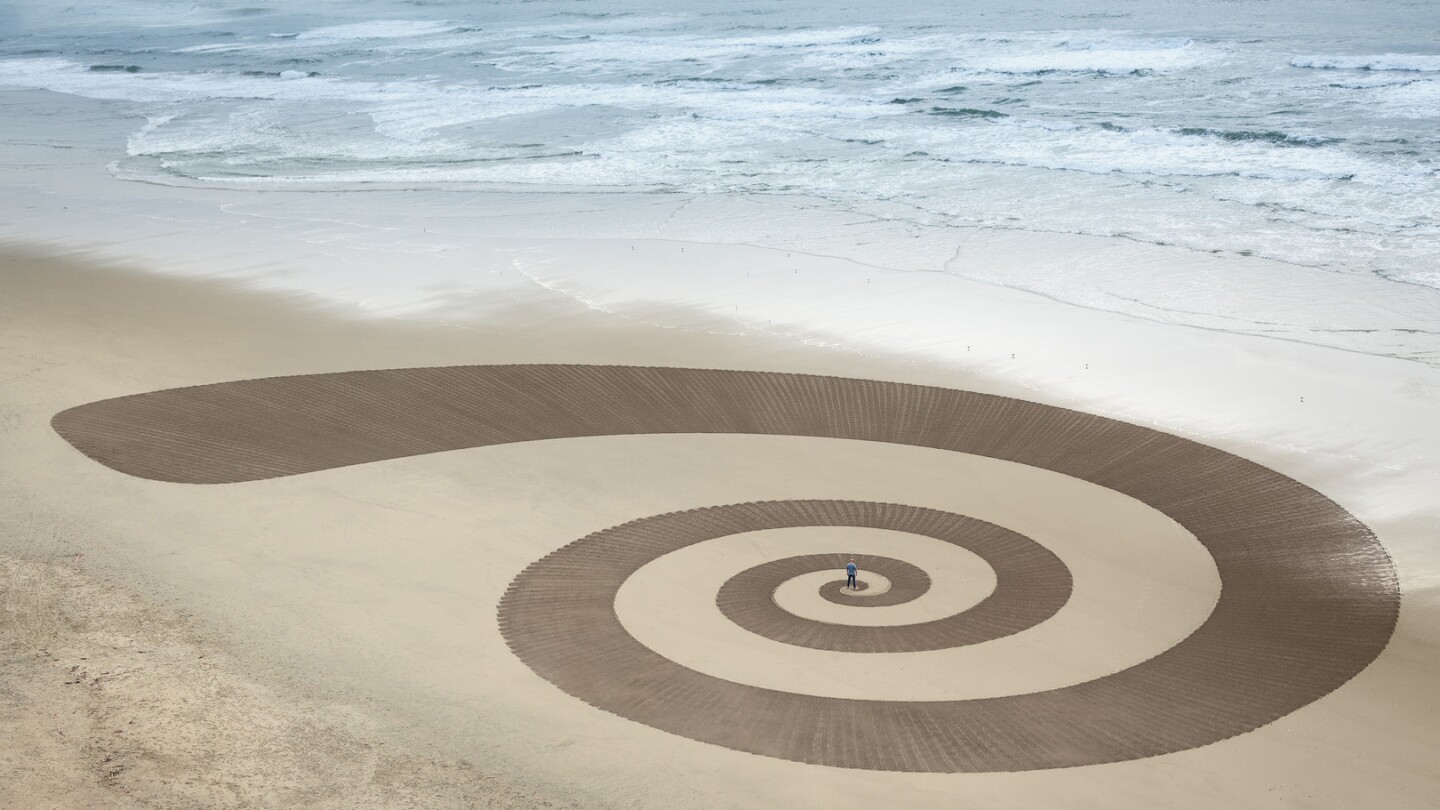 An aerial view captures a massive spiral design drawn onto sand. Beyond is a shoreline, slowly lapping at the coastline and erasing parts of the design. For scale, a man stands in the middle of the spiral, a mere speck in relationship to the greater artwork.