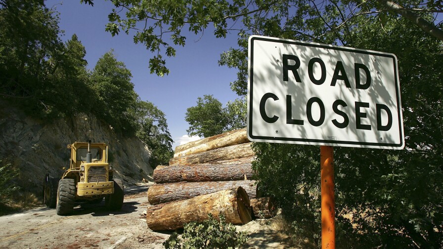 "Road closed" sign sits in the foreground while logs are stacked next to the closed road.