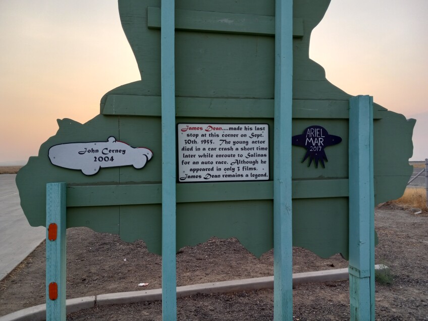 A sign on the back of a James Dean plywood mural reads, "James Dean made his last stop at this corner on Sept. 30th, 1955. The young actor died in a car crash a short time later while enroute to Salinas for an auto race. Although he appeared in only 3 films. James Dean remains a legend. A smaller sign to the left reads, "Jean Cerney, 2004" on a sign shaped like a car. A smaller sign on the right reads, "Ariel Mar, 2017," on a sign shaped like a UFO.