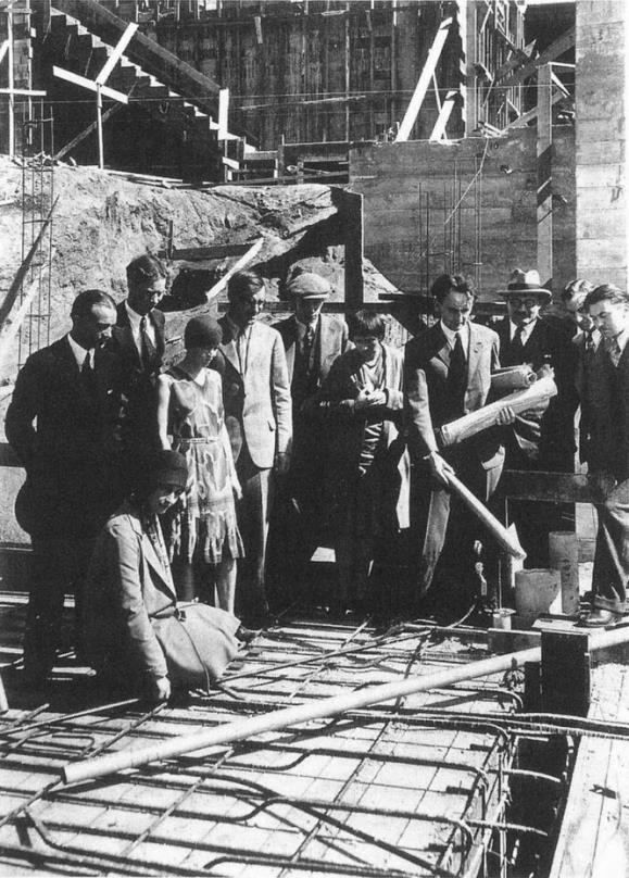 An archival black and white photo depicts a group of people huddled over reinforcing bars laid on the ground. A man in the group is gesturing at one of the bars with a roll of paper. The photograph is from 1929, so everyone is dressed in 1930s American fashion.