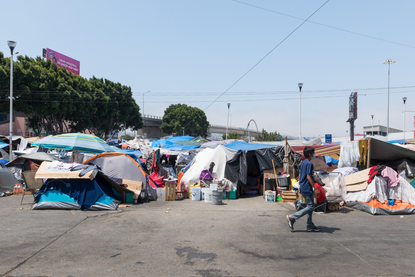 An asphalt surface is covered in various encampments. A boy is walking across the frame.