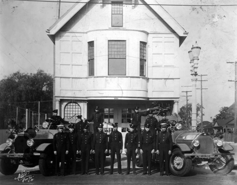 A black and white photo of a group of Black firefighters standing outside a firehouse with their firetrucks