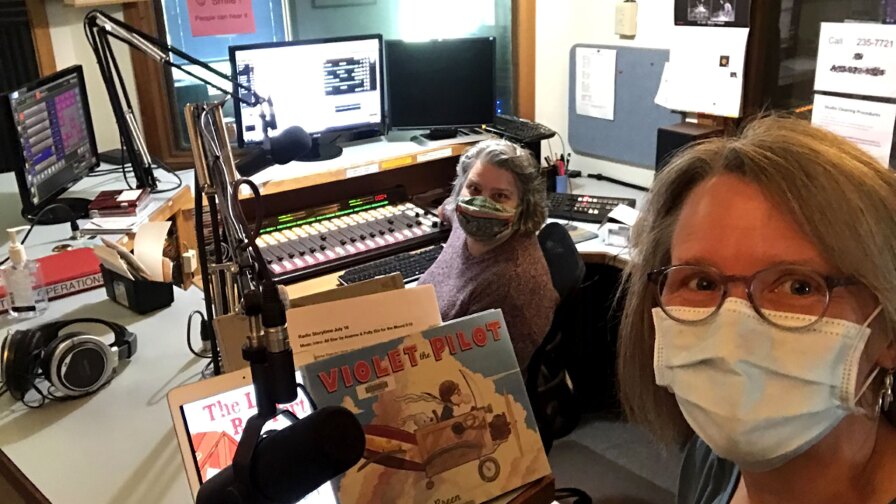 Two women wearing masks sit inside a radio recording studio