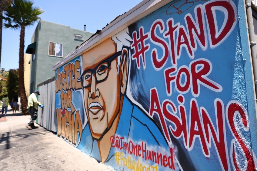 A colorful spray-painted mural adorns a low wall on the side of a building, depicting an Asian man with white hair, a white mustache and black glasses and the words "JUSTICE FOR VICHA" and "#STANDFORASIANS"