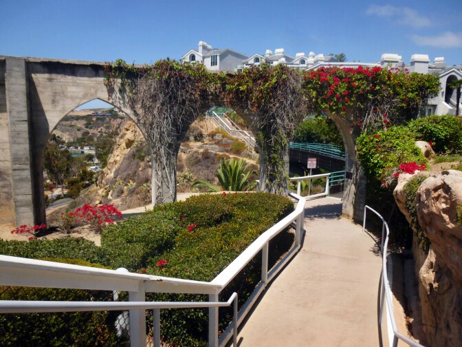 A paved walkway leads to a set of three stone arches covered in vining plants. The plants have bright green leaves and vibrant pink-red flowers.