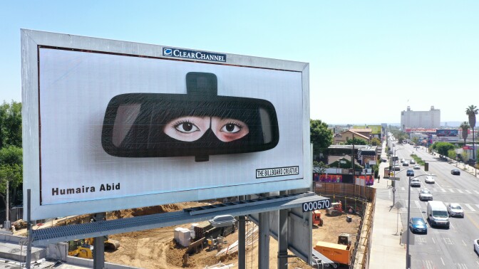 A billboard in L.A. shows a woman's reflection in a car's rear view mirror. Wearing a niqab, the woman's eyes are the only visible part of her face, and one eye bears a noticeable bruise. Below is a busy street with cars bustling by.