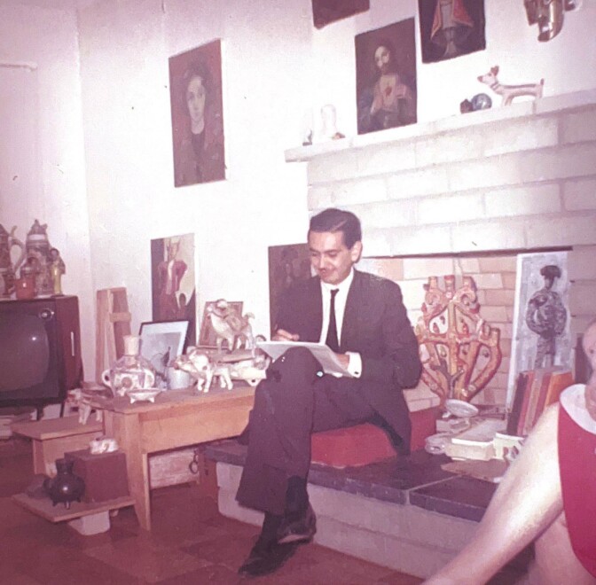 J. Sergio O'Cadiz Moctezuma wearing a black suit and tie, sitting on a fireplace mantle. His leg is crossed over the other and a writing surface is resting on his knee. He's looking down and appears to be writing something down. He's smiling.