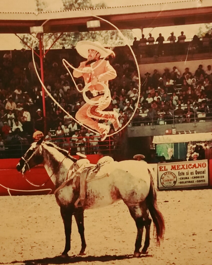 An old, yellowing photo of a young Esteban Escobedo jumping over a horse as a lasso of rope spins around his body. He is performing in a dirt arena where several spectators watch on in stands behind him.