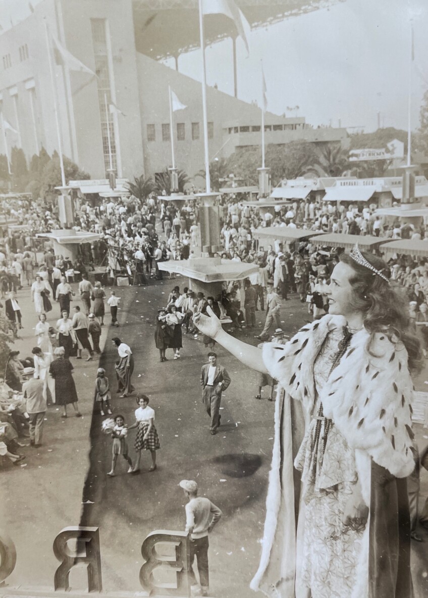A black and white photo of a young white woman standing above the L.A. County fairgrounds. She is wearing a gown, a royal cape and a crown atop her head. Below, attendees look up at her.