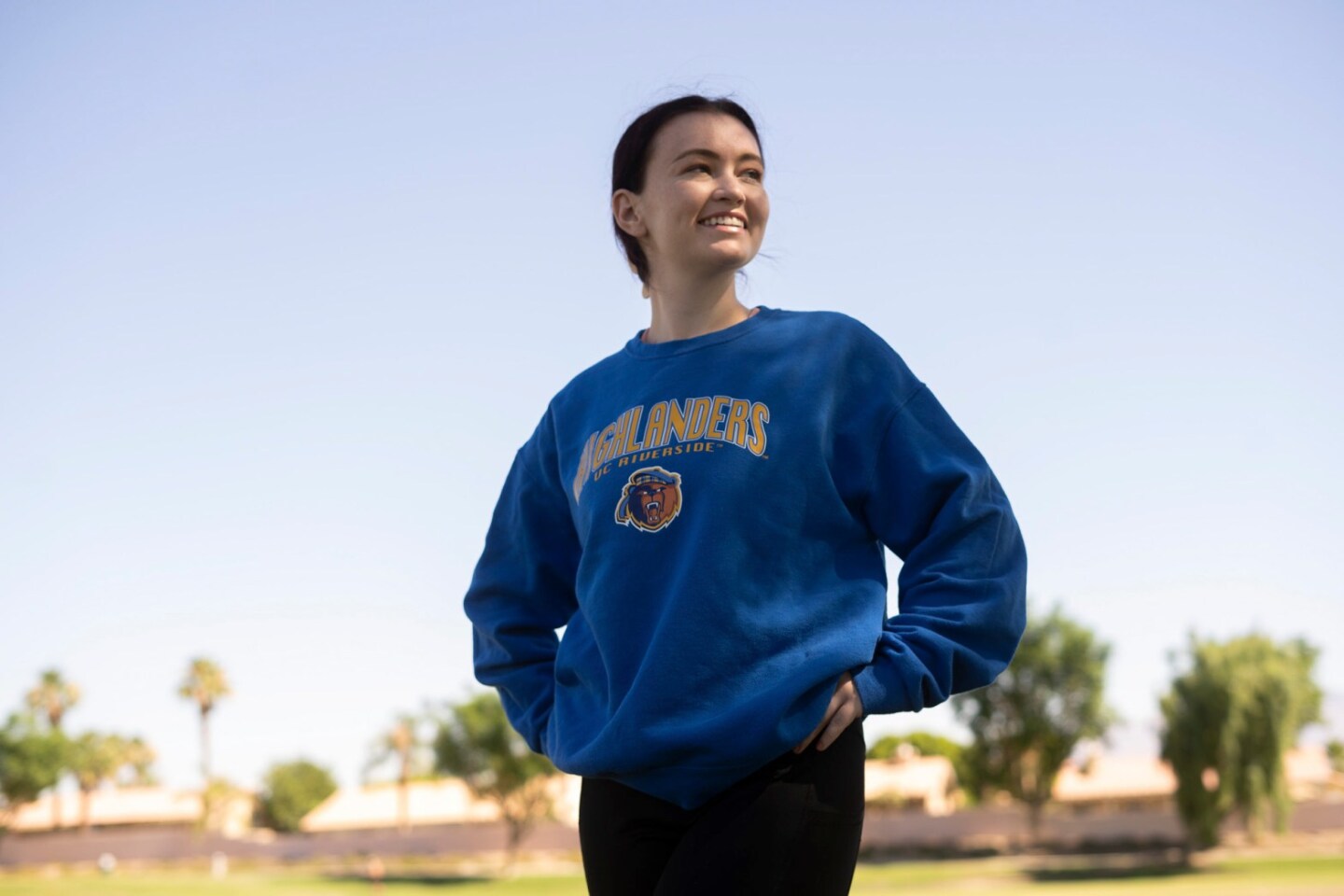 A young person wearing a blue sweatshirt and a long ponytail stands outside and looks to their left.