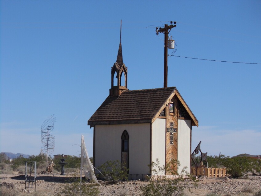 A lone chapel sits in the middle of a desert. Metal sculptures are strewn across the grounds surrounding the chapel. 