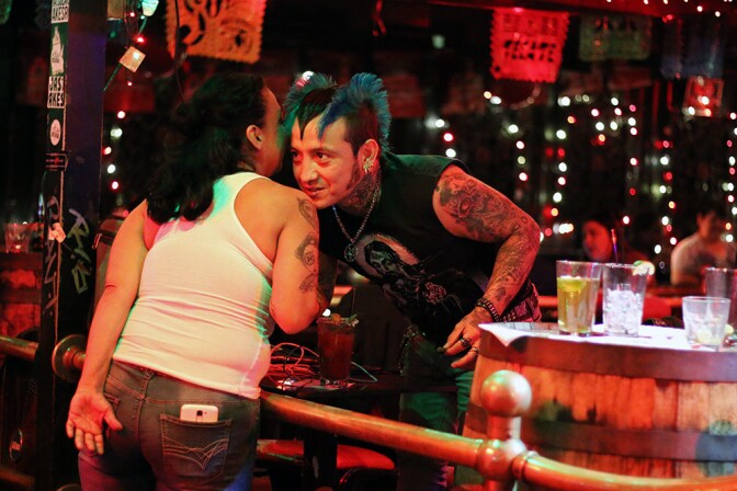 A woman whispers into the ear of a man with a skull tattoo and blue hair at La Cita.