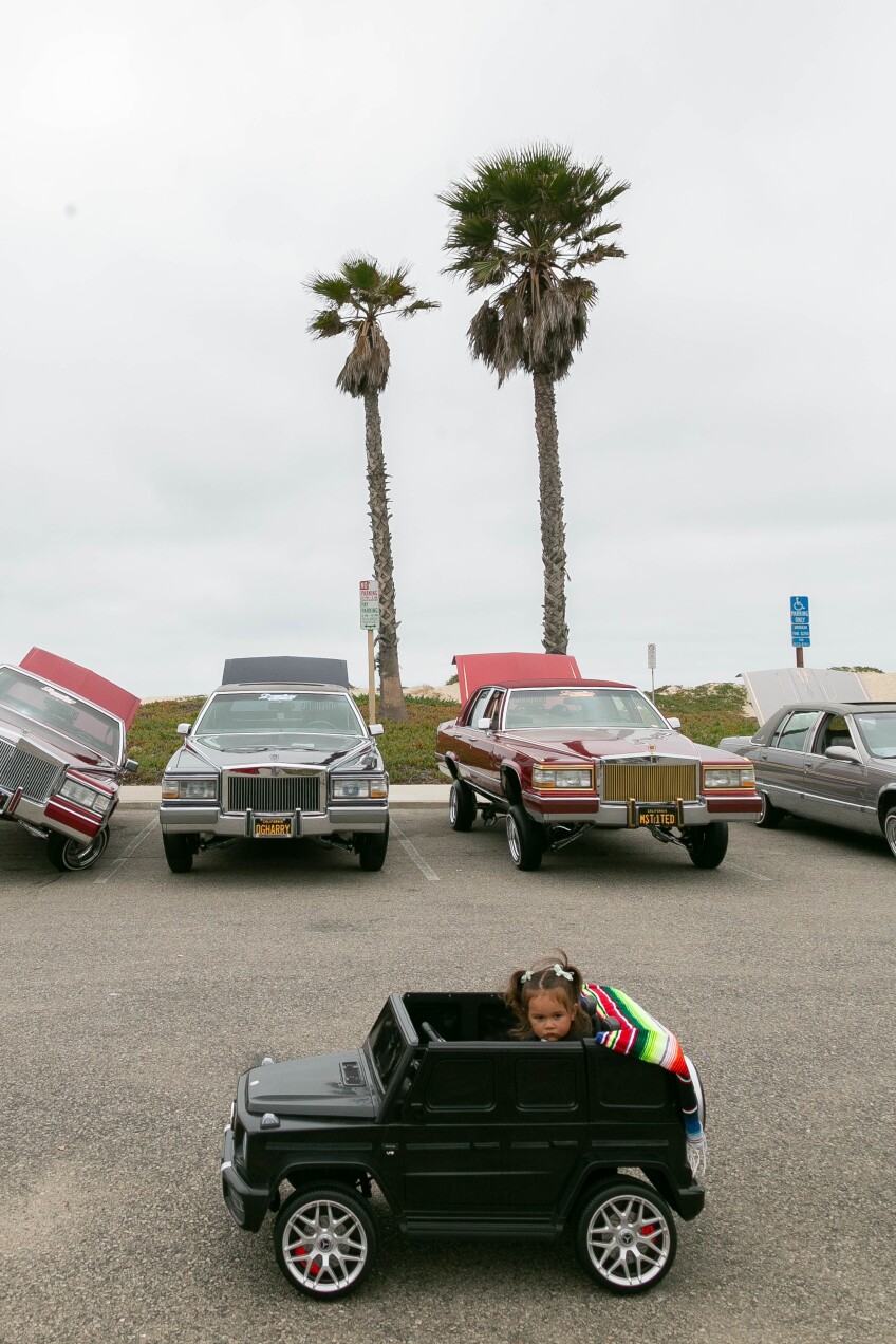 A little girl in a toy car behind low riders.