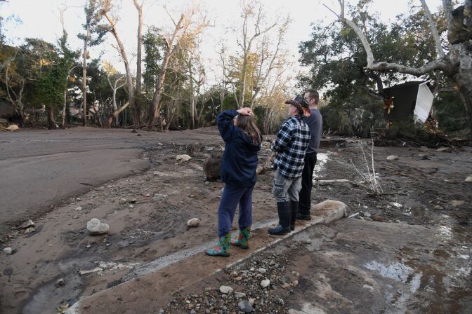 Three people stand looking at the destroyed landscape after a mudflow, one woman has her hands on her head