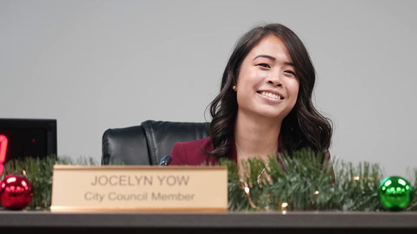 A woman smiles behind a nameplate on a desk that reads "Jocelyn Yow, City Council Member."