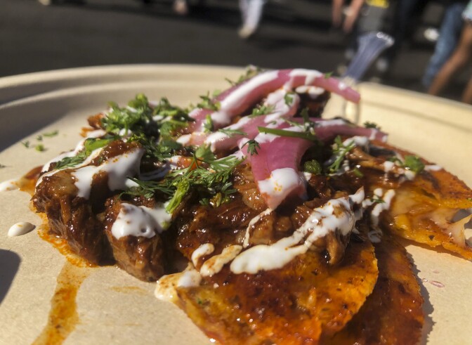 A close-up photo of goat birria tacos on a paper plate. The birria taco is smothered in a orange-red sauce. The entire dish is topped with pickled red onions and green herbs, possibly cilantro or parsley.