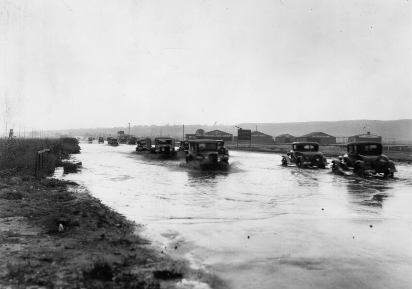 Photograph of flooded street looking south on Crenshaw from Exposition, January 1930. In the foreground at right, a wide street can be seen beside a patch of dirt and vegetation at left. A large layer of water covers the road. At center, at least seven cars trudge through the water. Large waves churn beneath the tires of the automobiles. In the extreme background at center, additional automobiles can be seen. To the right, a row of small buildings are visible.