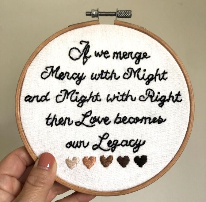 A hand-held embroidery hoop with white fabric stretched across with the words, "If we merge mercy with might and might with right, then love becomes our legacy," embroidered in black thread. The words are a quote from Amanda Gorman's inaugural speech. At the bottom of the embroidery hoop design are five flesh-toned embroidered hearts in different shades going from lightest to darkest.