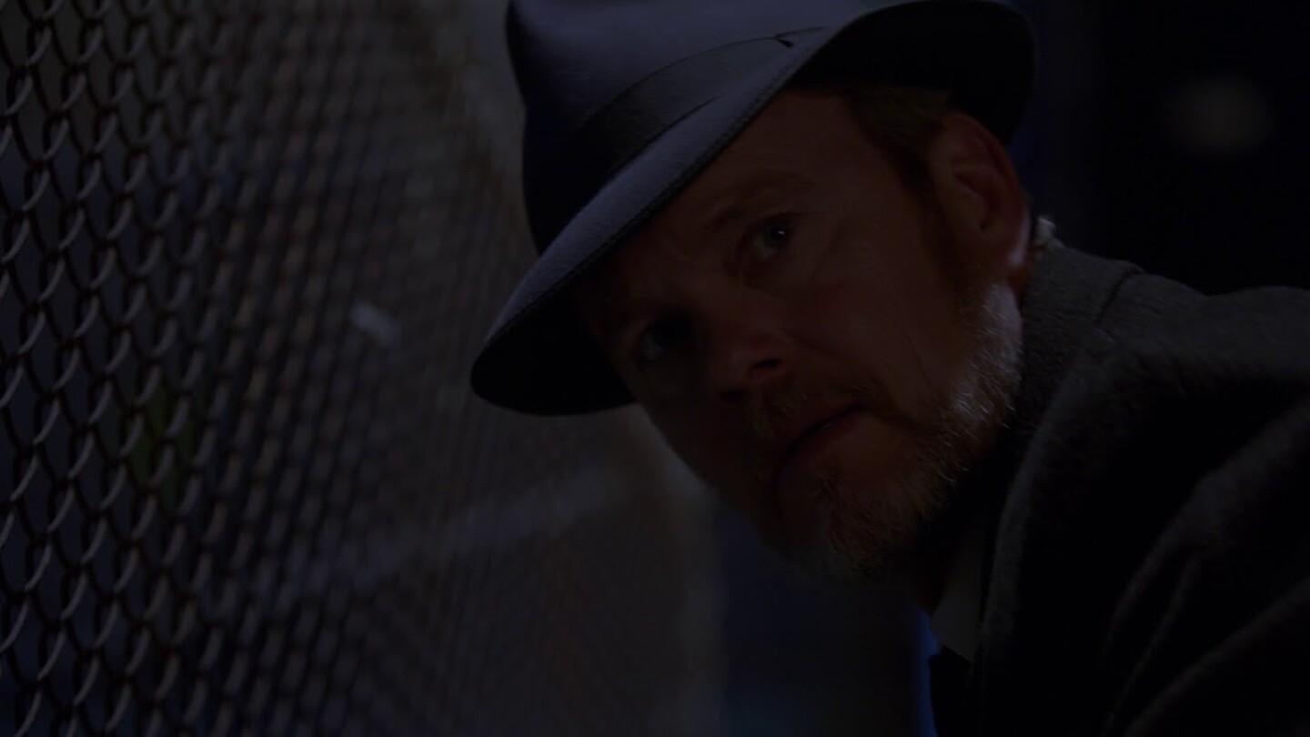 Craig Mclachlan wearing a hat and jacket next to a chain link fence in The Doctor Blake Mysteries.