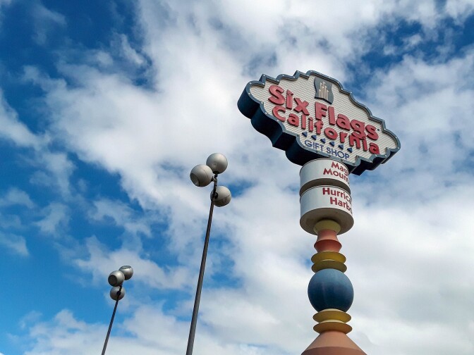 A sign for the Six Flags Magic Mountain gift shop. The sign is decorative and reads, "Six Flags California" with arrows pointing to the left and directly beneath lettering that reads, "Gift Shop." Below the sign are two panels. The top panel reads, "Magic Mountain" and the one below reads, "Hurricane Harbor." The sign is helped up with a decorative, uniquely shaped post and stands against a blue sky and fluffy white clouds.