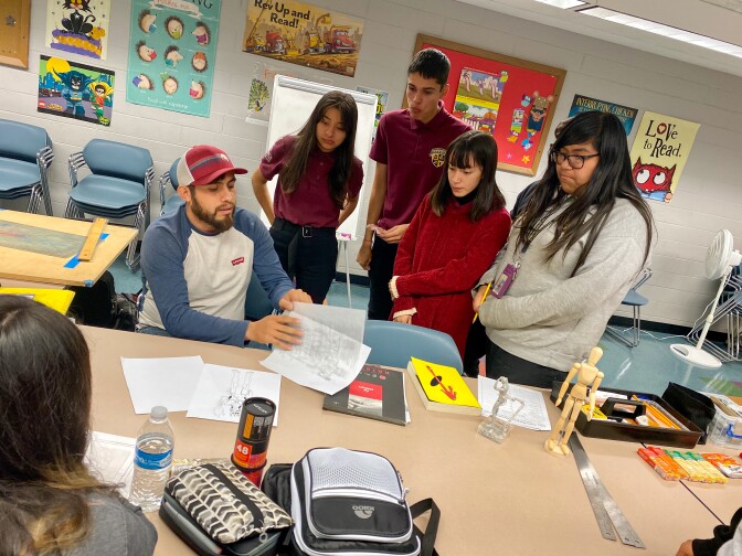 Artist Abraham Jarmillo guides a workshop about comic book coloring at a table in the Vernon - Leon H. Washington Jr. Memorial Branch Library. Students gather around him.