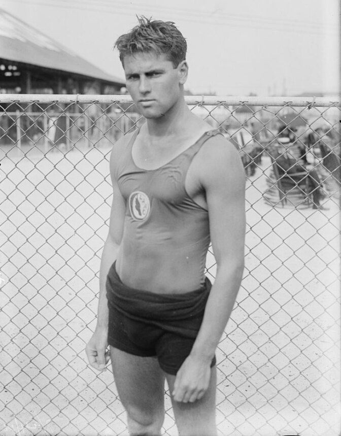 An old black and white photo of a young Tom Blake standing in front of a chain-link fence. He is wearing a men's bathing suit, still damp from the water. He has a serious expression on his face as he looks direct to camera.