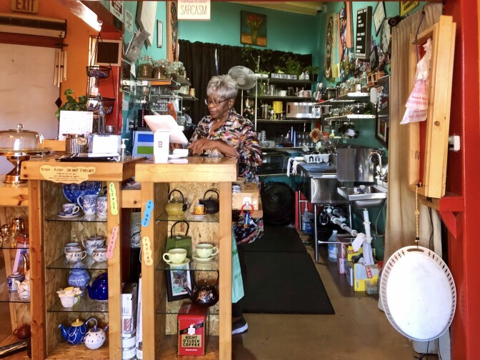 An older woman sits behind a counter. She is surrounded by an eclectic environment. The wall behind her is painted in a vibrant teal color. Mugs and teacups of various sizes, shapes and patterns are arranged in a shelf nearby.