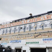 A three-store building with various attractions and stores painted on like "cafeteria," "groceria," "botanicas," and "mariachis." Above all is big orange lettering that reads, "Bienvenidos al Mercadito." Below that is a mariachi band painted on the building.