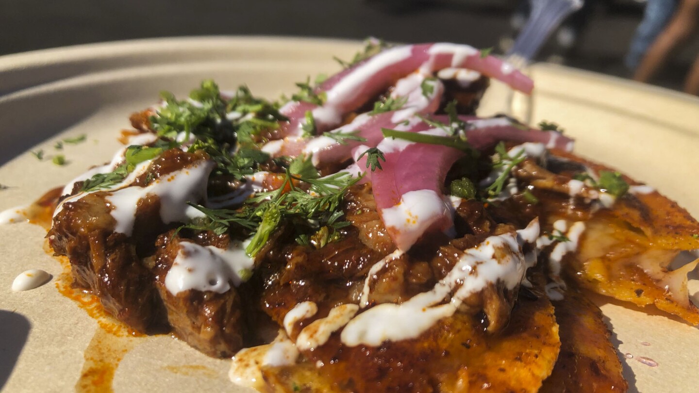 A close-up photo of goat birria tacos on a paper plate. The birria taco is smothered in a orange-red sauce. The entire dish is topped with pickled red onions and green herbs, possibly cilantro or parsley.