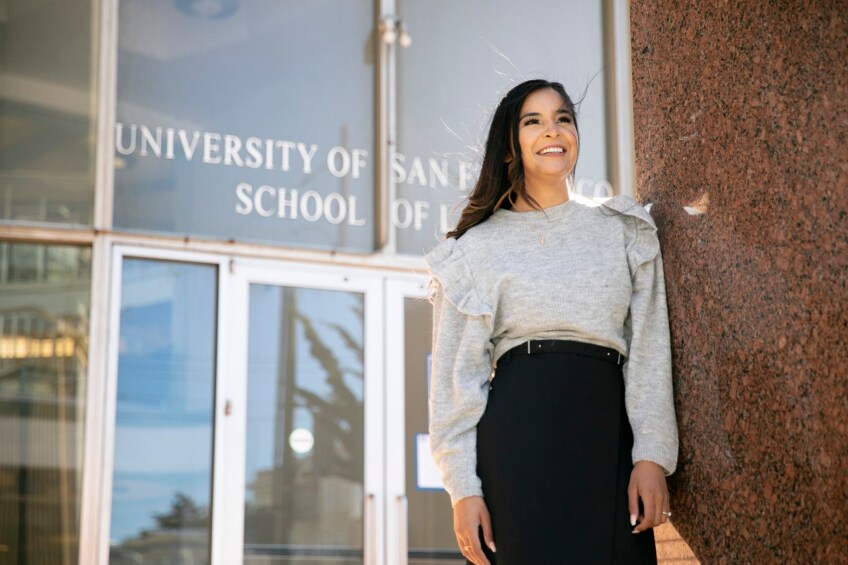 Second year law student Manjinder Kaur stands for a portrait at the University of San Francisco Law Library on May 24, 2021. Her long hair moves with the wind and she smiles. She is wearing a ruffled grey sweater and black bottoms.