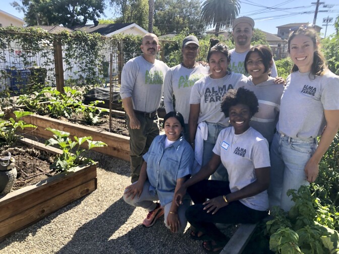 A group of people wearing shirts that read, "Alma Backyard Farms" pose for a picture. There are eight people posed in three rows. Two women are kneeling at the front, five people stand behind them and a taller man stands behind the whole group in a third row by himself. The group is surrounded by gardening beds filled with various leafy, green produce.