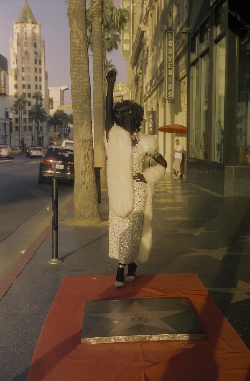 Muxxxe, adorned in a black morph suit under a sparkling white dress, curled wig and bedazzled jewelry, stands on a red carpet next to their Hollywood Walk of Fame star recreation. They're standing with one hand on their hip and the other in the air.