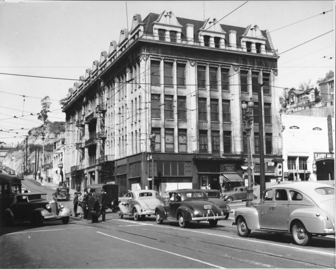 An archival black and white photo of a building with several windows. On the street that runs by it, vintage cars drive by.