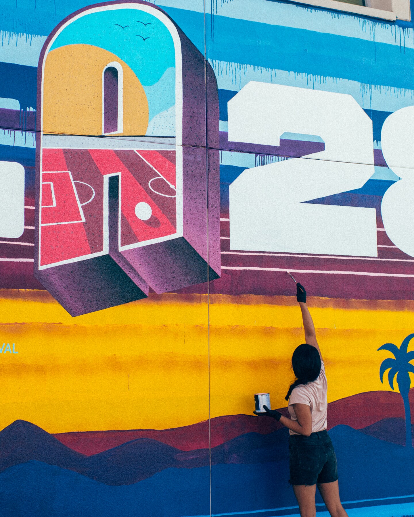 A young woman paints a a mural that says "LA28" with a sunset and palm tree silhouettes