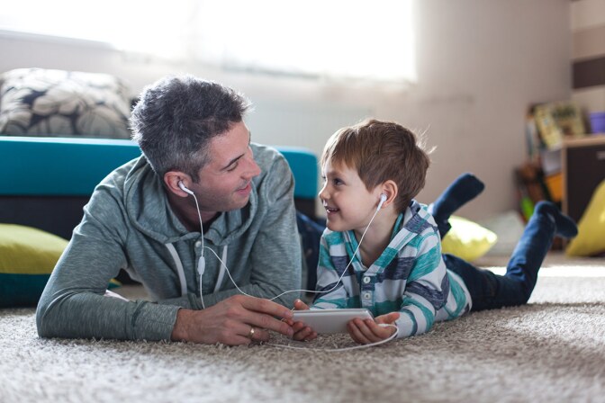 A man and a little boy watch something on a tablet together as they lie down on a carpet next to each other