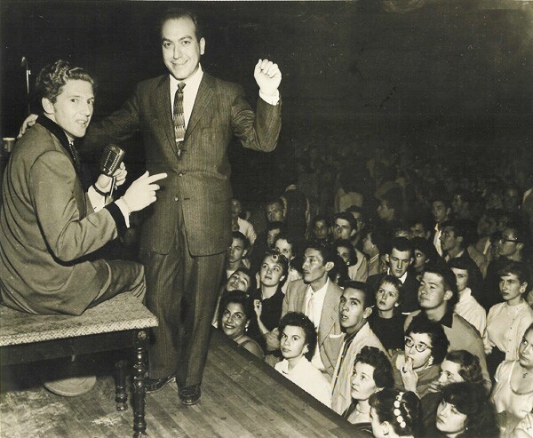 A black and white archival photo of two men on stage: Jerry Lee Lewis, who is sitting on a chair and turning back at the camera, and Art Laboe, who is standing with a fist in the air. Behind them is a crowd of concertgoers, looking up at them. 