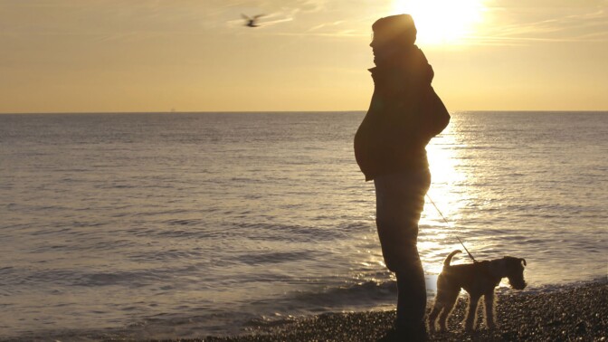 A pregnant transgender man looks pensively toward the sea while holding his dog on a leash. | From "Seahorse: The Dad Who Gave Birth"