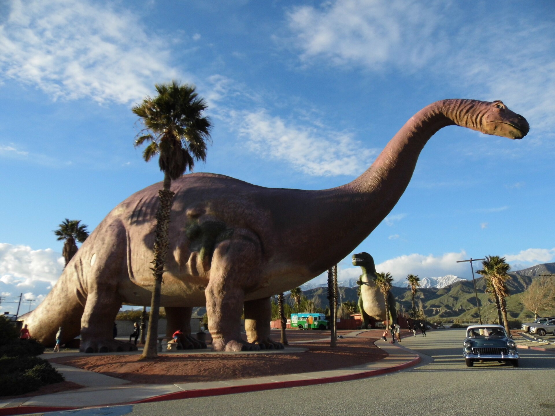 A large apatosaurus dinosaur sculpture with a long neck and tail towers over a road where a car drives by. In the background, a mountain landscape is visible and a tyrannosaurus rex sculpture. 