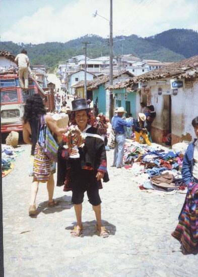Robert Lopez on a 1980 trip to Guatemala. He's standing in the middle of a cobble-stoned street wearing a black top hat and holding an art piece.