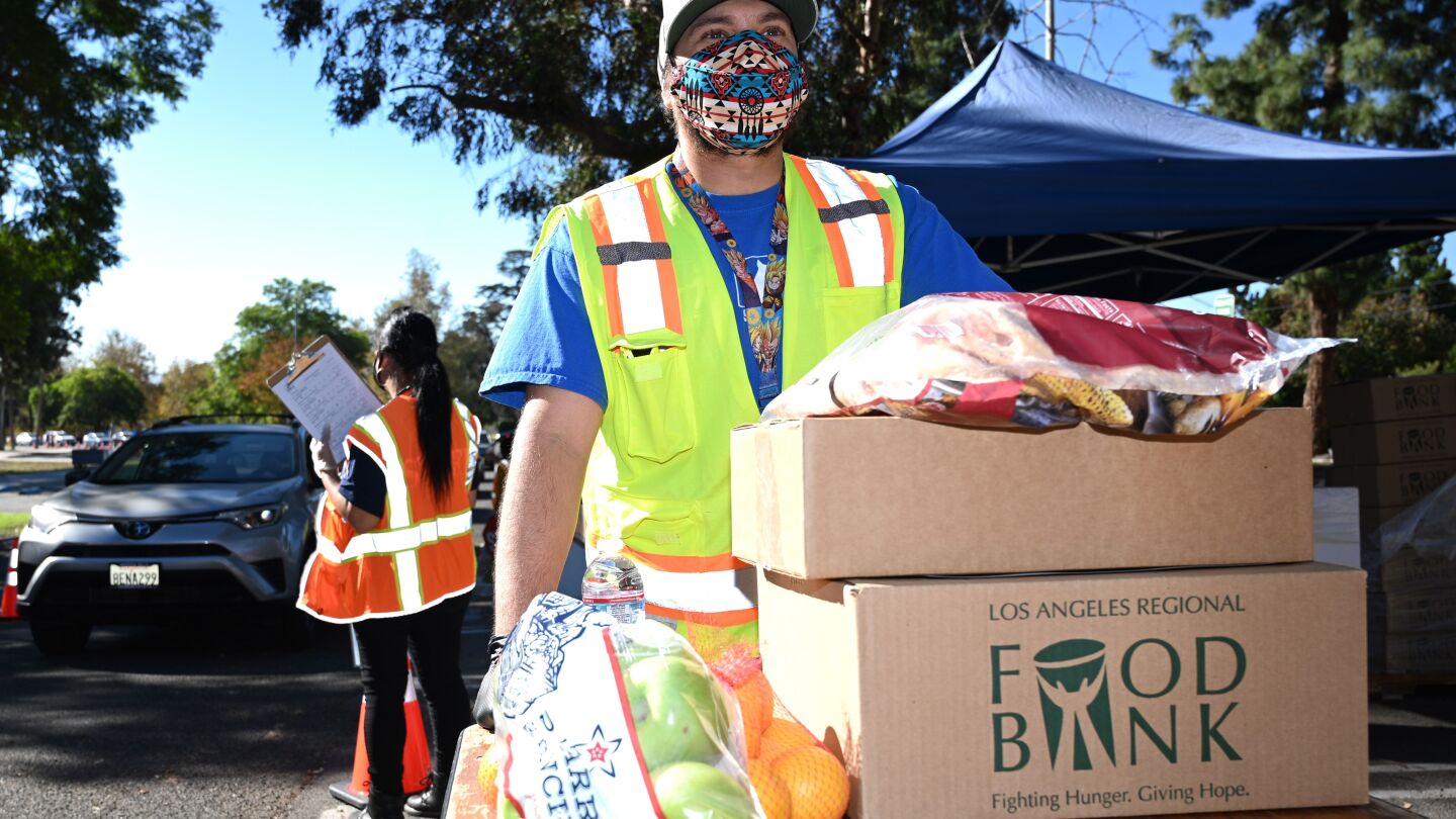 A man in a bright yellow reflective safety vest stands behind two boxes stacked on a table. There are also bags of fruit. The man is wearing a mask and looking off to the distance. Behind him is a person wearing a bright orange safety vest and holding a clipboard. Their back is facing the camera and it appears as if they are guiding a line of cars.