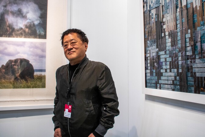 Taiji Terasaki stands with his hands tucked into his pockets and looking at the camera. He's wearing a black bomber jacket. Behind him are two of his art pieces. The art piece to the left are still printed photographs of the environment. The piece on the right is woven sheet metal with environmental photos printed onto the metal.