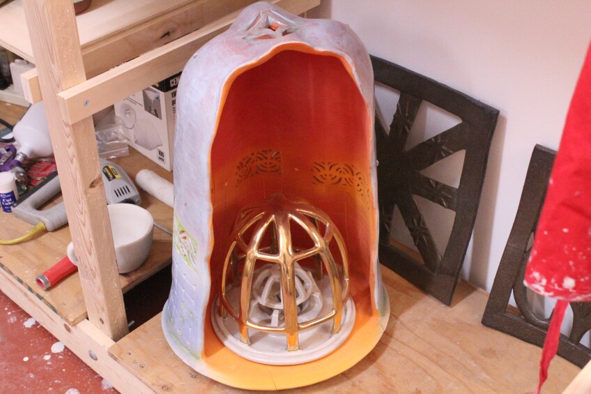 A bell-shaped ceramic sculpture appears to be cut in half. Inside, is a gold dome frame. The walls of the bell interior are orange with geometric shapes cut out of it. The exterior is a grayish-purple. 